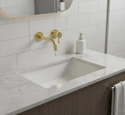 Gouden, aan de muur gemonteerde badkamerkraan boven een rechthoekige, witte wastafel die is ingebouwd in een marmeren aanrechtblad. Op de achtergrond is een witte, rechthoekige tegelmuur en een verlichte, ronde spiegel te zien