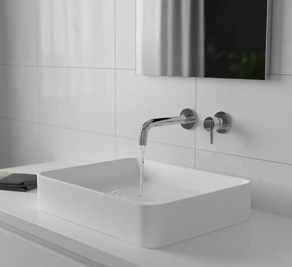 Close-up van een moderne, wandgemonteerde chromen kraan boven een rechthoekige witte opbouwspoelbak in een badkamer met witte marmeren tegels.