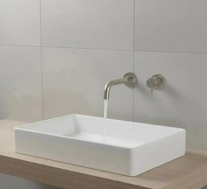 Een strakke, moderne badkameropstelling met een witte rechthoekige opbouwspoelbak die rust op een licht houten aanrechtblad. Boven de spoelbak stroomt water uit een geborsteld nikkel of mat roestvrijstalen wandgemonteerde kraan. De muur achter de spoelbak is bedekt met grote, lichtgrijze, vierkante tegels.